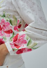 Blusa bianca con un motivo floreale caratterizzato da fiori rosa e rossi. Maniche lunghe con polsi arricciati e delicati dettagli a intaglio.