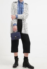 Vit fluffig cardigan över en blå denimskjorta, i kombination med svarta kapade byxor och svarta snörstövlar. Håller en liten marinblå handväska.