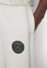 Lys grå joggerbukser lavet af blødt stof, med et sort cirkulært logo der har teksten "PARIS" og "SAINT-GERMAIN", accentueret med beige syninger.