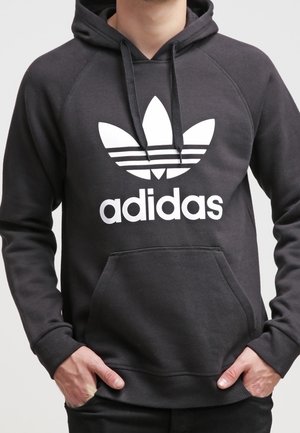 Person iført en sort Adidas-hoodie med hvidt trefoil-logo og tekst, hænderne i forlommen, stående mod en ensartet baggrund.