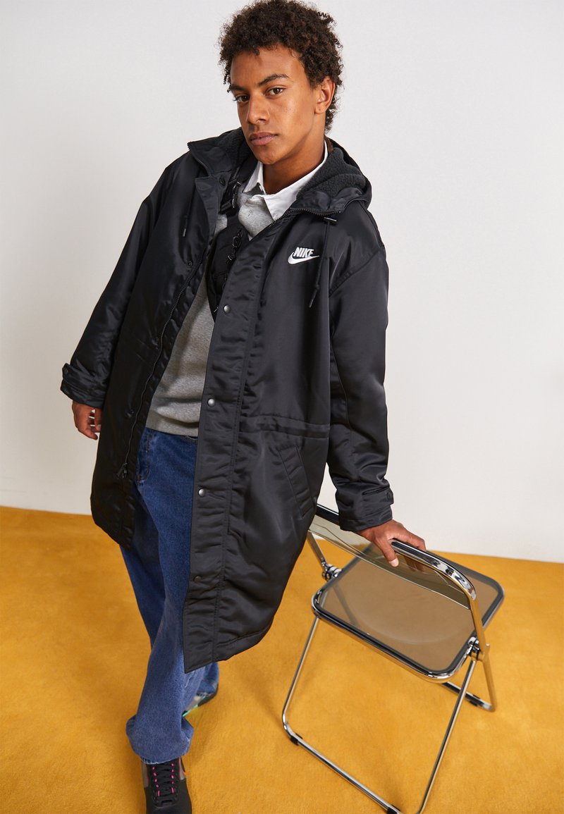 Manteau long en nylon noir avec capuche, présentant une fermeture éclair et des boutons, des poches à l'avant et un logo Nike sur la poitrine. Jeans bleus et chaussures noires.