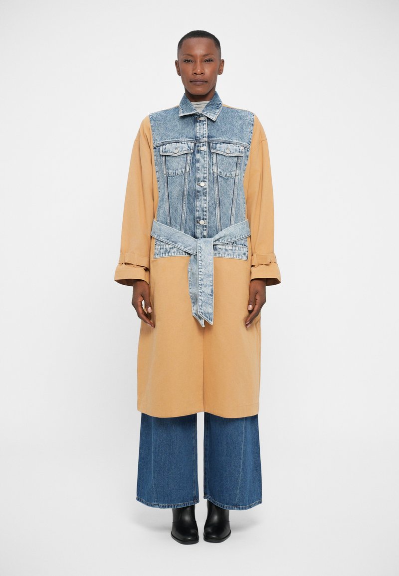 Robe chemise en denim et beige, présentant un devant à boutons, une ceinture à la taille et des manches longues. Pantalons en denim évasés larges et bottines noires.