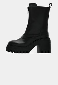 Black leather ankle boot with chunky lug sole, block heel, side zipper, and pull tab at the back on plain white background.