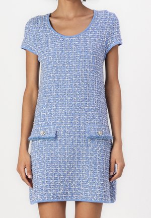 Kvinde iført en kortærmet, knælængde blå tweedkjole med tekstureret ternet mønster og to forlommer med klap og dekorative knapper.