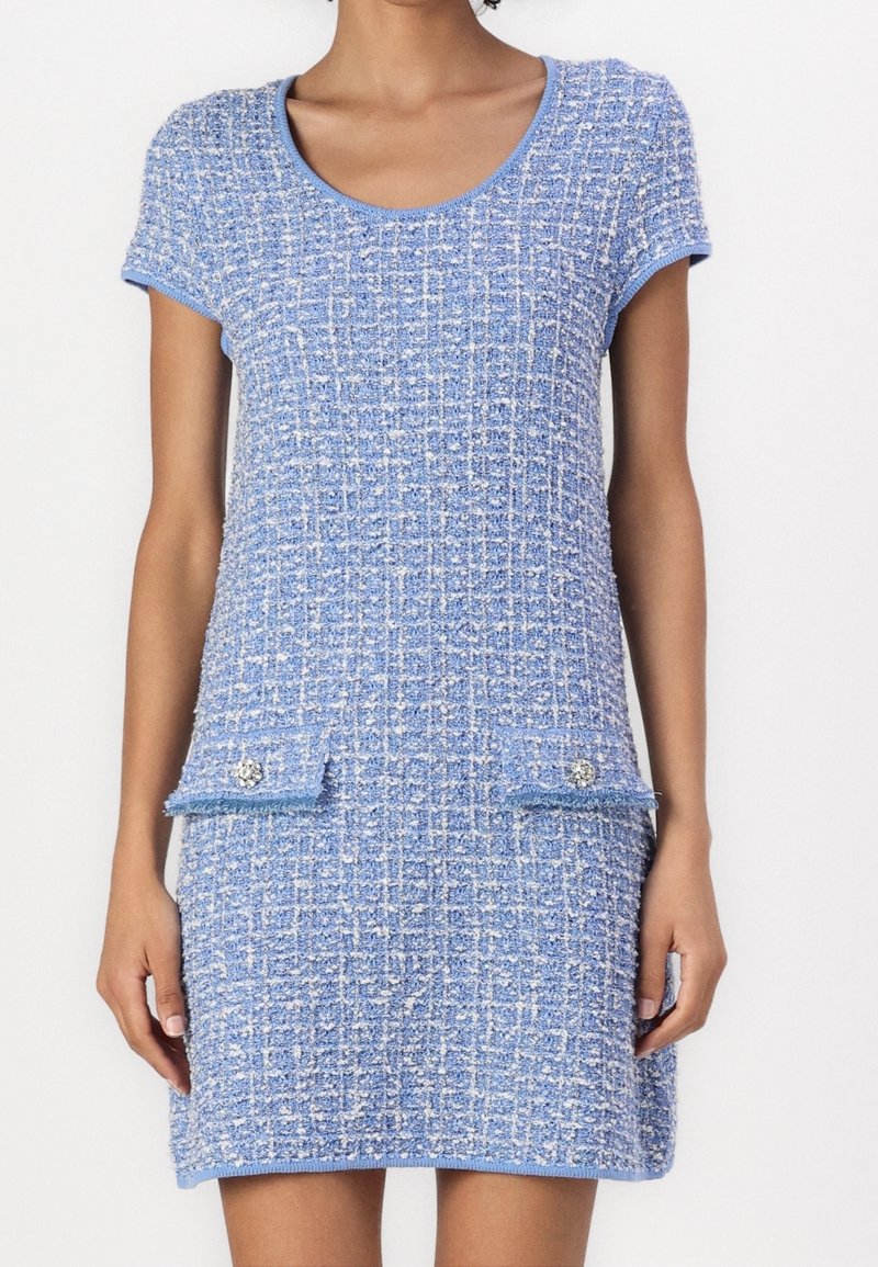 Femme portant une robe en tweed bleue à manches courtes, longueur genou, avec un motif à carreaux texturé et deux poches avant à rabat ornées de boutons décoratifs.