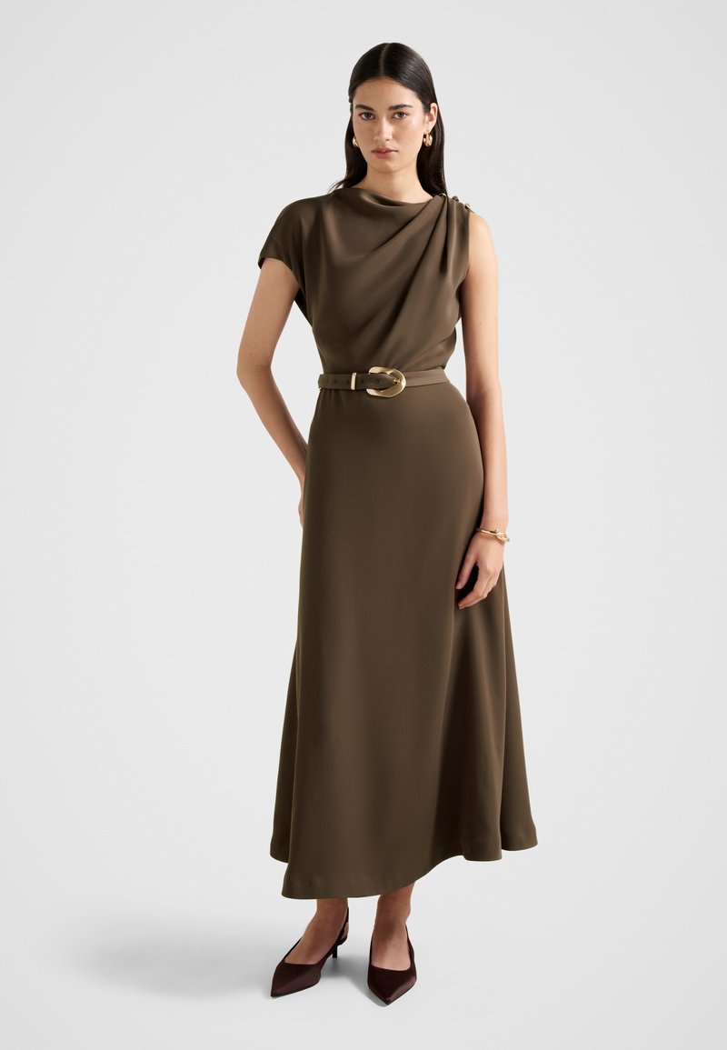 Γυναίκα με ελαιόχρωμο, αμάνικο φόρεμα με ζώνη και πιετά λαιμόκοψη, χρυσούς κρίκους στα αυτιά, βραχιόλι και σκούρα παπούτσια με μυτερά άκρα, πάνω σε λευκό φόντο.