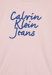 Jasnoróżowa bluza z niebieskim haftem z napisem "Calvin Klein Jeans" w swobodnej czcionce. Gładka, miękka struktura.