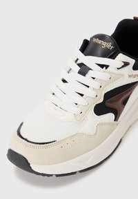 Sportssko med en hvit mesh-overdel, beige semsket skinn detaljer, svarte striper og en brun logo. Har hvite skolisser og en teksturert svart såle.