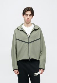 Zielona krótka kurtka z przednim zamkiem błyskawicznym, kapturem i czarnymi paskami akcentowymi. Połączona z czarnymi spodniami z logo. Gładka faktura.