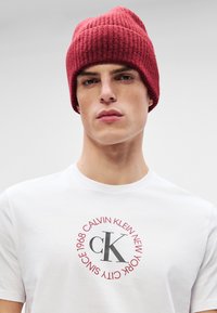 Gorro de malha vermelho com textura canelada. Usado com uma t-shirt branca com um logótipo circular cinza e vermelho contendo o texto "Calvin Klein New York".