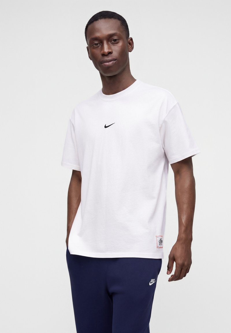 Mand iført en hvid Nike T-shirt og navyblå Nike bukser, stående mod en ensartet lys baggrund.