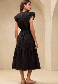 Robe noire à longueur genou avec un col froncé, une taille élastique et un ourlet en couches. Présente un motif floral et des détails en œillets.