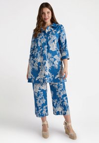 Blå blommig tunika och byxset med vita tropiska mönster, trekvartsärmar och krage, kombinerat med beige kilklackssandaler.