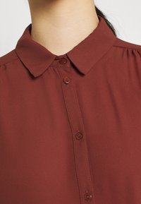 Blouse bordeaux à col avec une texture soyeuse, présentant une patte de boutonnage et un léger froncé aux épaules.