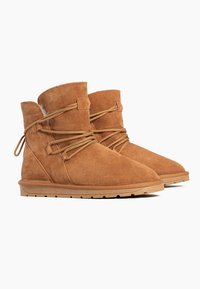 Beige Wildleder-Ankle-Boots mit runder Zehenpartie, die mit überkreuzten Schnürsenkeln und einem kurzen, weichen Kragen ausgestattet sind. Flache Gummisohle für Komfort.