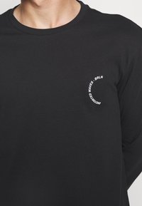 Schwarzes Langarm-T-Shirt aus Baumwolle mit einem kleinen weißen, kreisförmigen Grafikdesign mit Text auf der linken Brustseite. Einfaches Design.