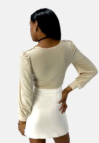 Beige top met lange mouwen en geplooide schouderdetails, gecombineerd met een witte rok. De stof oogt glad en aansluitend, wat een strakke uitstraling geeft.