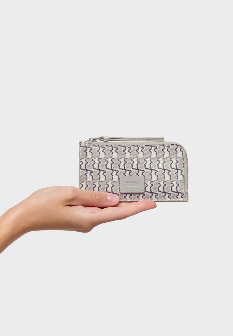 Gray fabric wallet with purple wavy pattern, zipper closure, and a rectangular metal logo patch. Hand holding the wallet for scale.