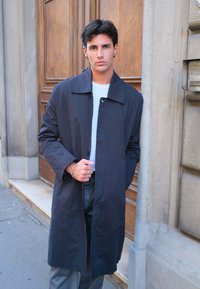 Ung mand med mørkt hår iført lang mørk frakke over hvid skjorte, stående ved en trædør og en stensfacade udendørs.