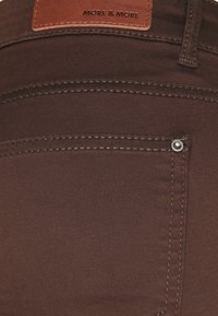 Brown denim fabric features a back pocket with visible stitching and a small metal rivet. A leather label is attached at the waistband.
