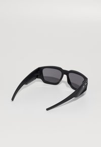 Gafas de sol negras con un diseño moderno y elegante, lentes ahumados grises y brazos curvados y estilizados que presentan un acabado texturizado y un detalle de logo.