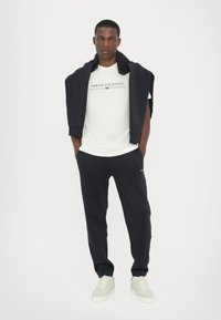 White t-shirt with "Armani Exchange 1991" print, paired with dark blue sweatpants and light-colored sneakers. Sweater draped over shoulders.