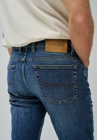 Vaqueros de mezclilla azul desvaído con un ajuste slim. Incluye un parche de logo de cuero marrón y bolsillos traseros estilizados con detalles de costura.
