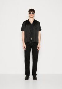 Camicia nera a maniche corte con pieghe verticali, abbinata a pantaloni e scarpe nere. Gli accessori includono occhiali da sole scuri e una fibbia dorata.