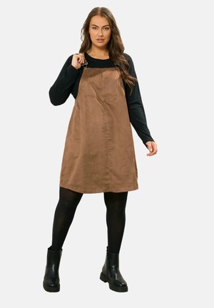 Bruine suede-effect tuinbroek jurk met verstelbare schouderbanden, gecombineerd met een zwart langemouwen top en zwarte enkellaarsjes.