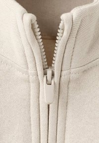 Kremasto barvna jopa z zadrgo, izdelana iz teksturirane pletene tkanine, s centralno zadrgo z belo zaponko in nežnimi šivi preko prsnega koša.