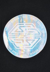 Kružna, hologramska oznaka s bijelim geometrijskim logotipom i natpisom "MISIÓN CONTROL - PUTUJTE IZA SVEMIRA - CSBL" na crnoj pozadini.