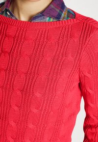 Röd stickad tröja med kabelmönster, rund halsringning och ribbad detalj vid fåll och muddar, bärs över en rutig skjorta.