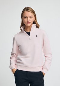 Ljusrosa zip-up sweatshirt med hög krage, mjukt tyg, ribbade ärmslut och en liten svart logotyp på vänster bröst.