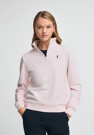Sudadera de cremallera en rosa claro con cuello alto, fabricada en un tejido suave, puños acanalados y un pequeño logotipo negro en el lado izquierdo del pecho.