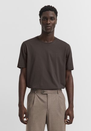 Mann trägt dunkelbraunes kurzärmeliges T-Shirt und beige hoch taillierte Hose und steht vor einem einfarbigen hellen Hintergrund.