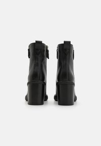 Botas de tobillo de cuero negro con un acabado texturizado, tacón alto de bloque, cierre de cremallera trasero y pequeñas correas decorativas a cada lado.
