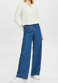 Camisa blanca de manga larga con cuello en V y rayas verticales azules, combinada con jeans de mezclilla azul de piernas anchas y zapatillas blancas. Fondo liso.