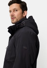 Czarna, odporna na warunki atmosferyczne kurtka z regulowanym kapturem, frontem na zamek i naszywką z logo na rękawie. Posiada matowe wykończenie i gładką teksturę.