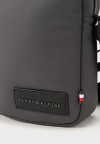 Szary plecak nylonowy z fakturowaną czarną metką z wytłoczonym napisem "TOMMY HILFIGER". Zawiera mały akcent w kolorach czerwonym, białym i niebieskim.