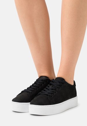 Une paire de baskets noires montantes avec des semelles blanches en plateforme portées pieds nus sur fond uni.