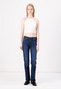 Cropped top blanco con tirantes finos, combinado con jeans rectos de color azul oscuro. Modelo vistiendo zapatos negros, de pie frente a un fondo neutro.