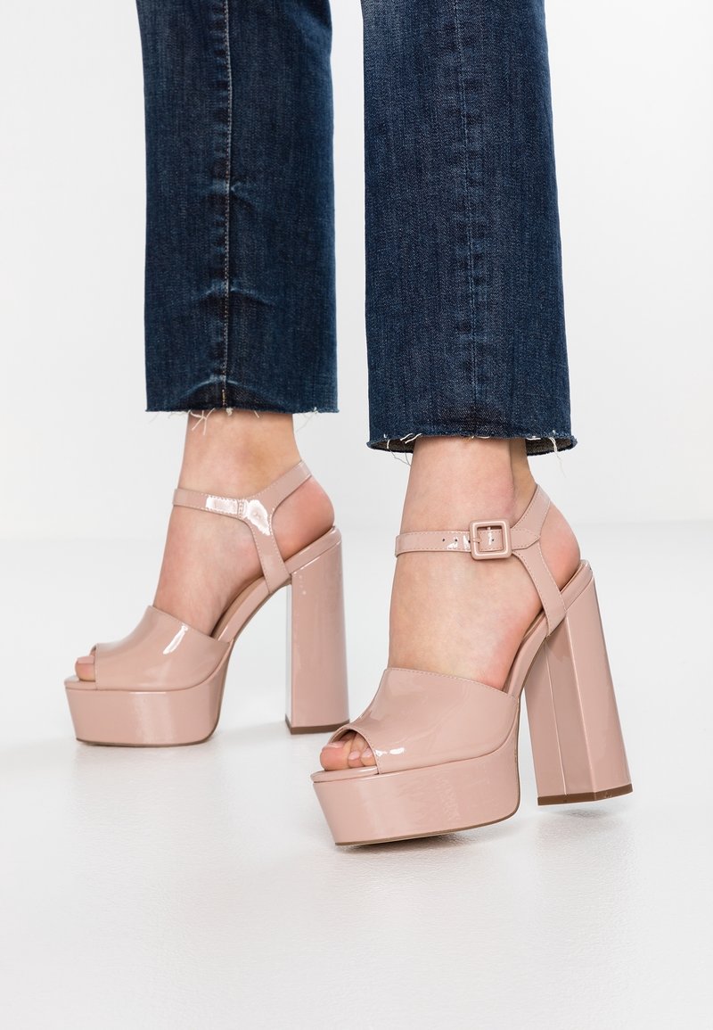 Pieds portant des sandales compensées à talons hauts vernis couleur nude avec des brides à la cheville et un jean court bleu foncé sur un fond uni.