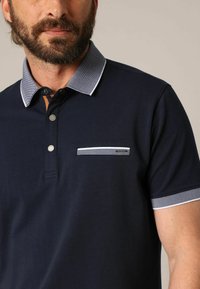 Polo shirt marine avec un col texturé et des accents bleu clair, trois boutons et une petite poche avec une bordure à motifs.