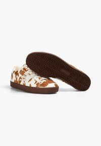 Zapatillas de lona blancas con patrones abstractos en marrón, punta redondeada y suela de goma marrón. Incluyen cordones planos y una suela texturizada.