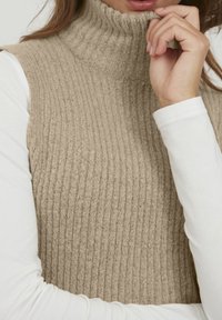 Nainen beigea ribbineulottu hihaton pooloneule valkoisen pitkähihainen paidan päällä, käsi lähellä leukaa ja huulet näkyvissä.