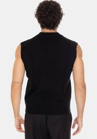 Uomo con capelli ricci che indossa un gilet nero senza maniche in maglia e pantaloni neri, in piedi di spalle con un tatuaggio sul braccio sinistro.