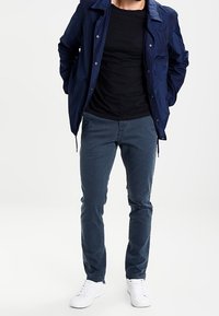 Blå nylonjacka med tryckknappar, svart t-shirt, blå byxor och vita sneakers. Avslappnad passform och minimalistisk design.