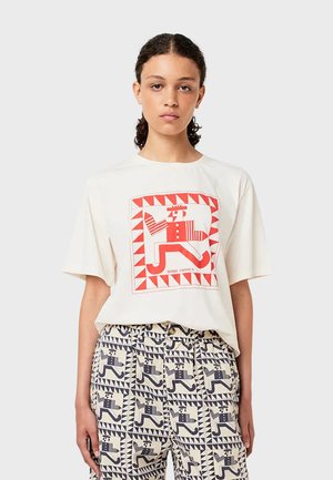 Krämfärgad t-shirt med kort ärm, prydd med ett röd- och vitfärgat geometriskt mönster, tillsammans med mönstrade byxor som har blå och krämfärgade designelement.