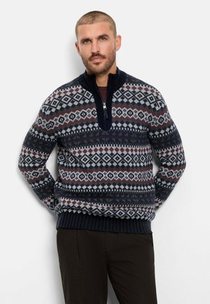 Homme aux cheveux courts et à la barbe portant un pull en maille à motifs bleu marine, blanc et bordeaux avec un col zippé partiel et un pantalon foncé.
