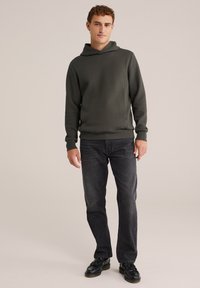 Temno zelena rebrasta kapuca z kengursko žepom in nastavljivim kapuco, v kombinaciji s temnimi jeans hlačami in črnimi čevlji.